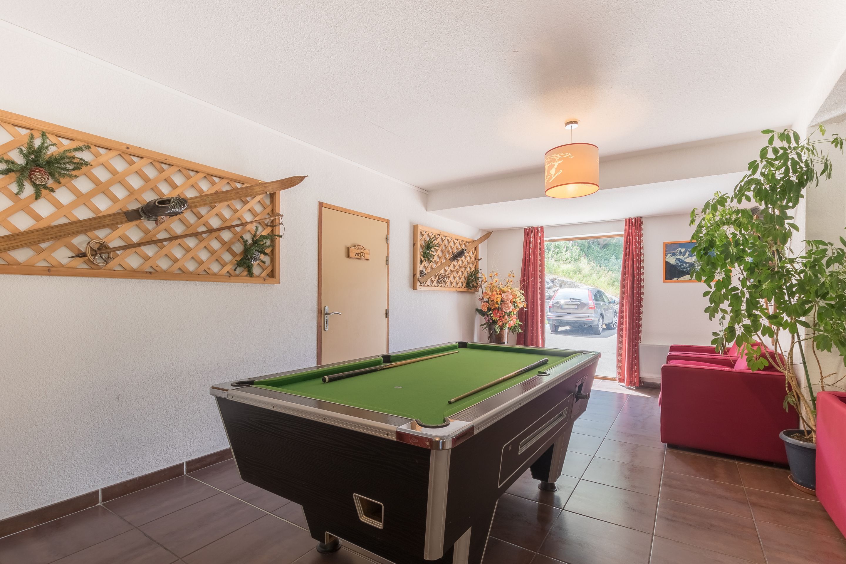 Une table de billard dans une pièce dont un mur est orné d’un treillage en bois, avec une porte portant l’inscription « VENT » et une plante en pot.