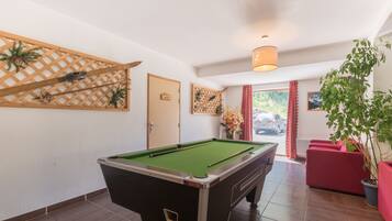 Une table de billard dans une pièce dont un mur est orné d’un treillage en bois, avec une porte portant l’inscription « VENT » et une plante en pot.