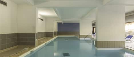 A swimming pool with a tiled floor, a changing room with a shower, and a view of the outside deck with lounge chairs.