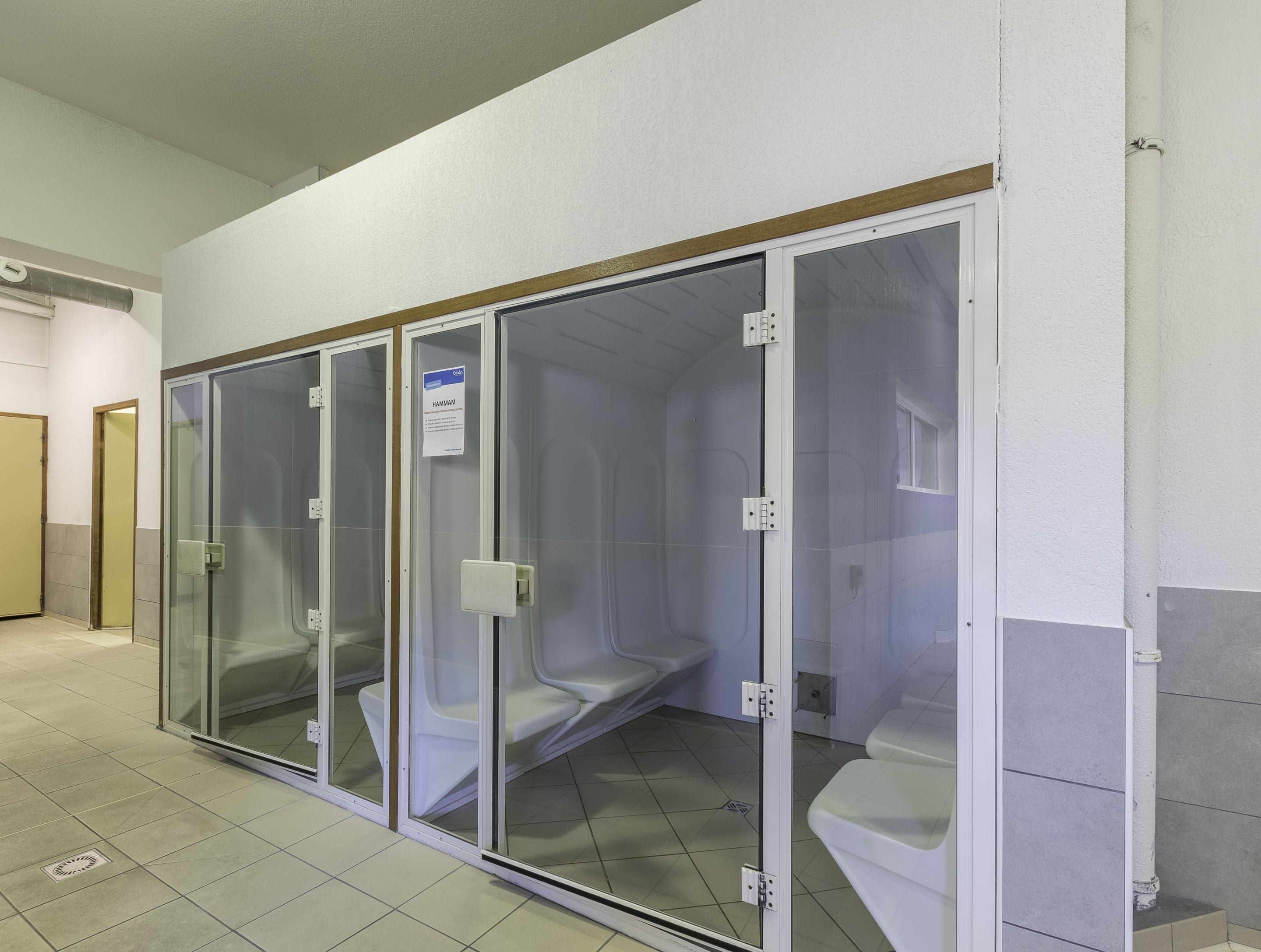 Een rij privé toiletcabines met glazen tussenschotten, een bordje met daarop Hamam en een betegelde vloer.