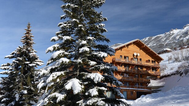 A wooden chalet with balconies, surrounded by snow-covered trees and mountains.