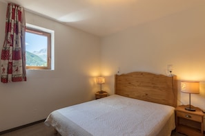 A bedroom with a wooden headboard, a single bed with a white quilt, two bedside tables with lamps, a window with curtains, and a view of mountains. - Résidence Odalys Pra Sainte Marie (Vars)