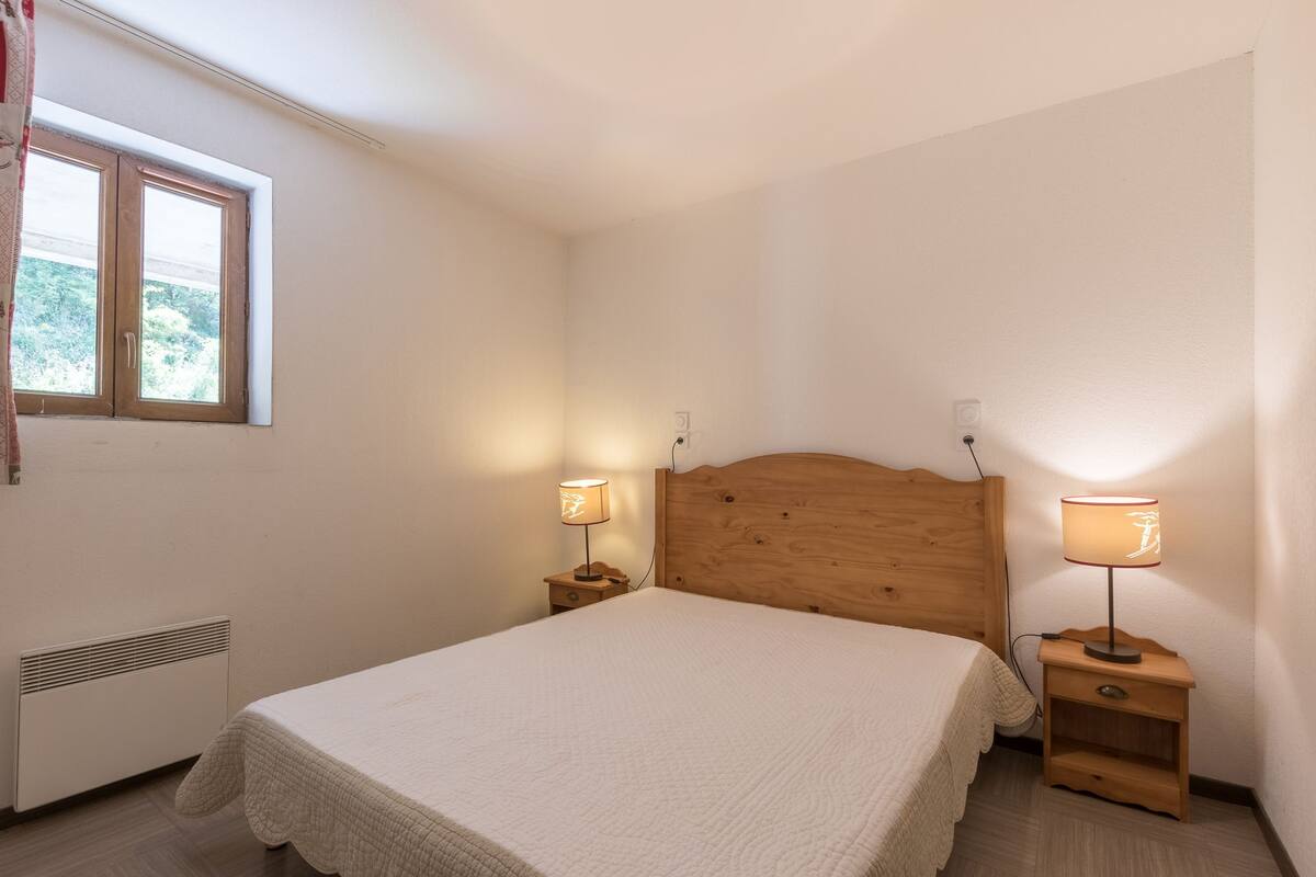 A bedroom with a wooden headboard, two bedside tables with lamps, a radiator, and a window with a view of greenery.