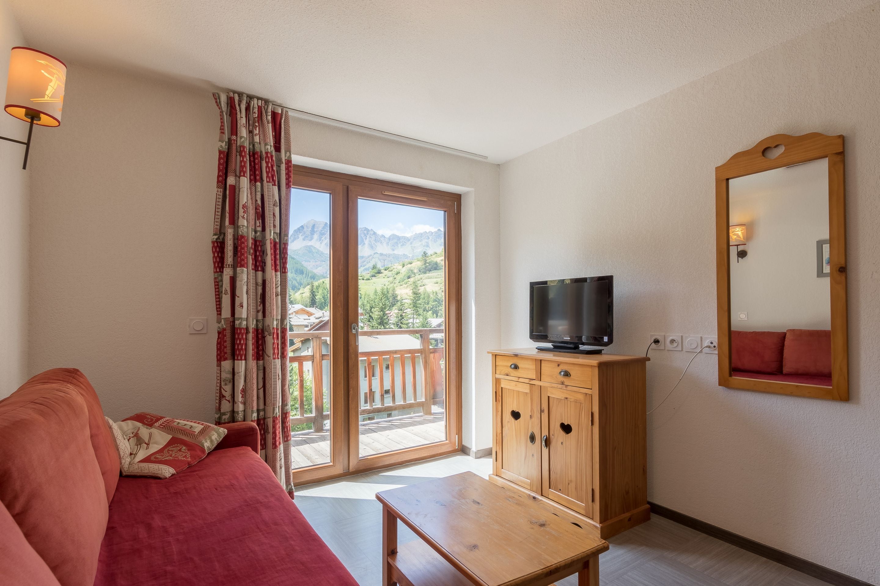 Una habitación con sofá rojo, una mesa de madera, un televisor sobre un mueble de madera y un ventanal con vistas a montañas.
