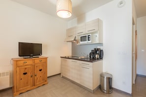A compact kitchen with a microwave, stove, and sink. There is a wooden cabinet with drawers and a television on top. A trash can is placed near the sink.