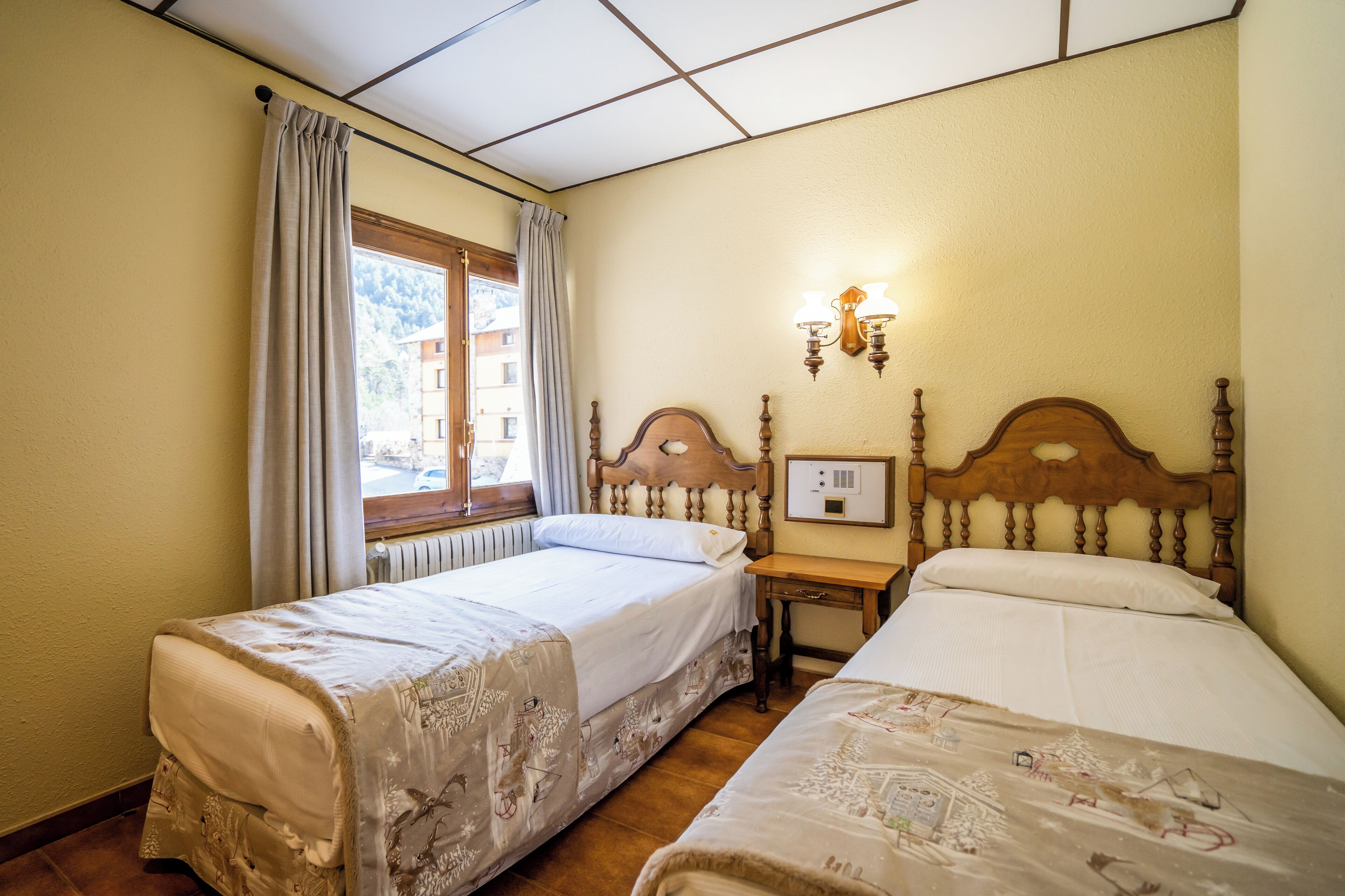 Habitación de hotel con dos camas individuales, cabecera de madera, mesita de noche, ventana con cortinas y lámpara fijada a la pared.