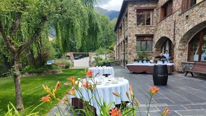 Un área de comedor al aire libre con mesas dispuestas para una comida, rodeada por un edificio de piedra y vegetación.