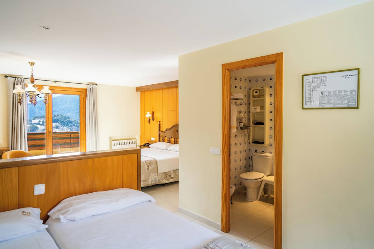 Une chambre d’hôtel avec deux lits, une tête de lit en bois, une fenêtre avec des rideaux, un lustre et une salle de bain visible à travers une porte ouverte.