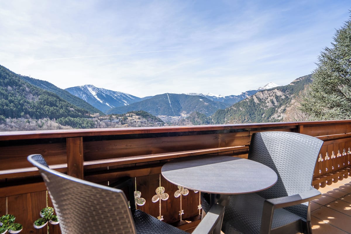 Une terrasse avec une table ronde et deux chaises, offrant une vue sur un paysage montagneux aux sommets enneigés et aux forêts denses.