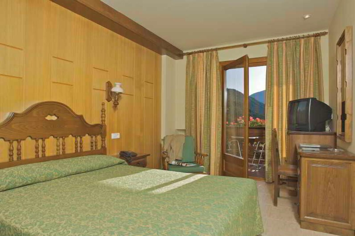 A hotel room with a wooden headboard, a bed with green bedding, a television, a desk, and a view of a mountainous landscape through the window.