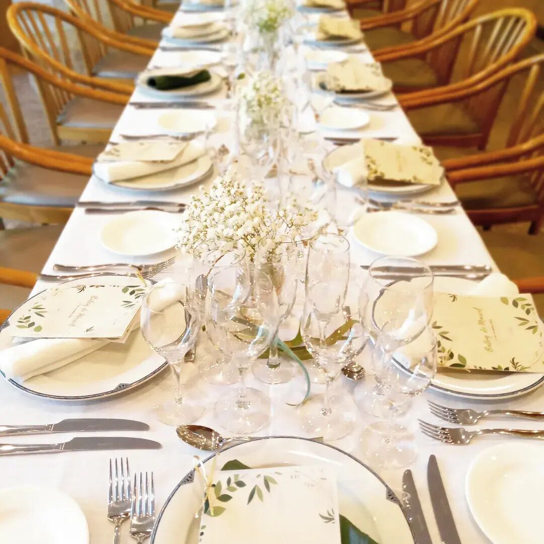Una larga mesa de comedor vestida con manteles blancos, cubiertos y cristalería, flanqueada por sillas de madera.