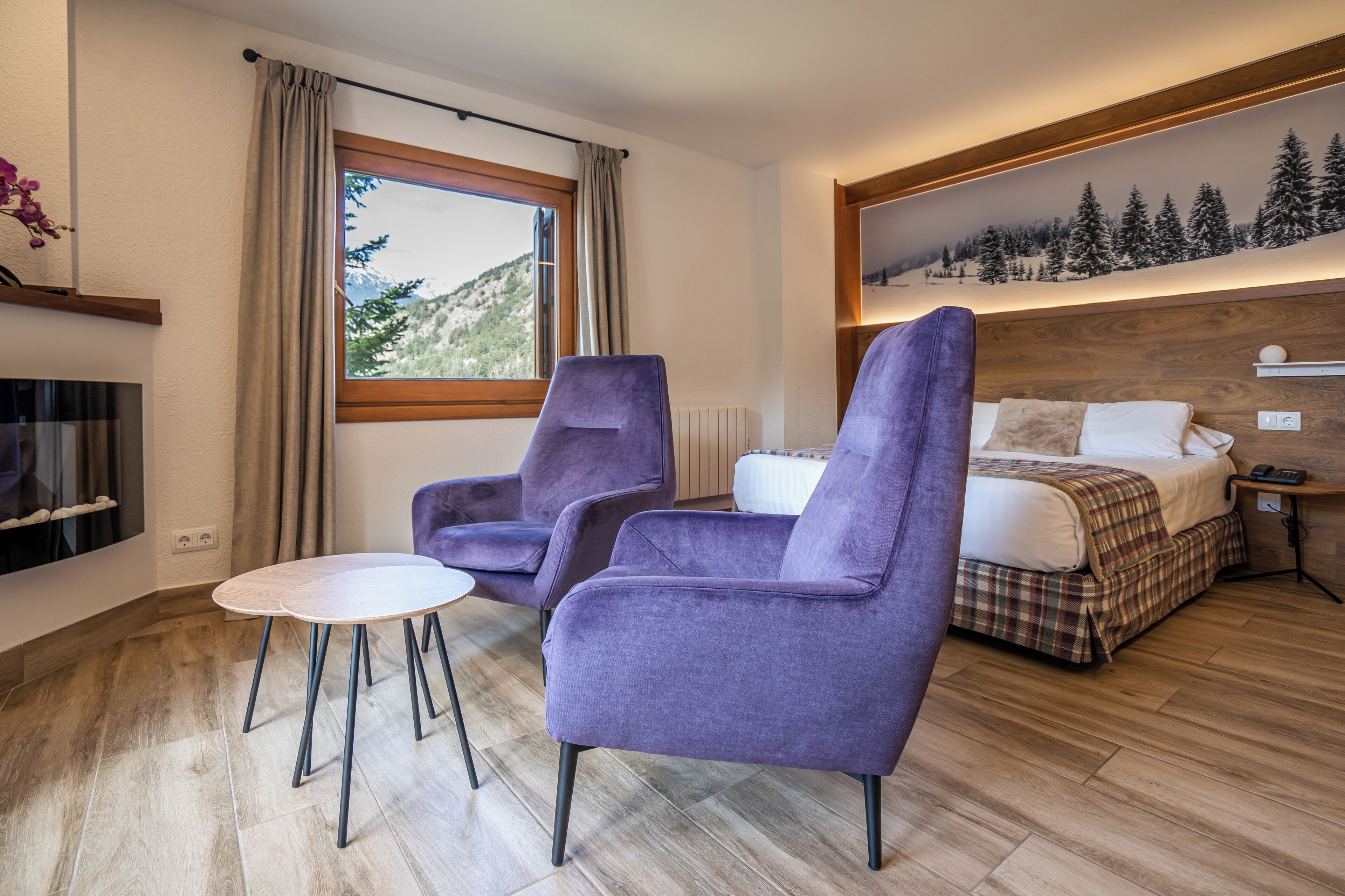 Una habitación de hotel moderna con una cama grande, dos sillones morados, una mesita y vistas a una montaña nevada a través de una ventana.
