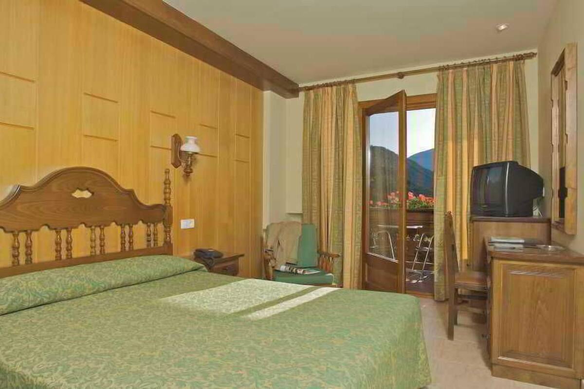 Une chambre d’hôtel avec une tête de lit en bois, un lit avec une literie verte, une télévision, un bureau et une vue sur un paysage montagneux par la fenêtre.