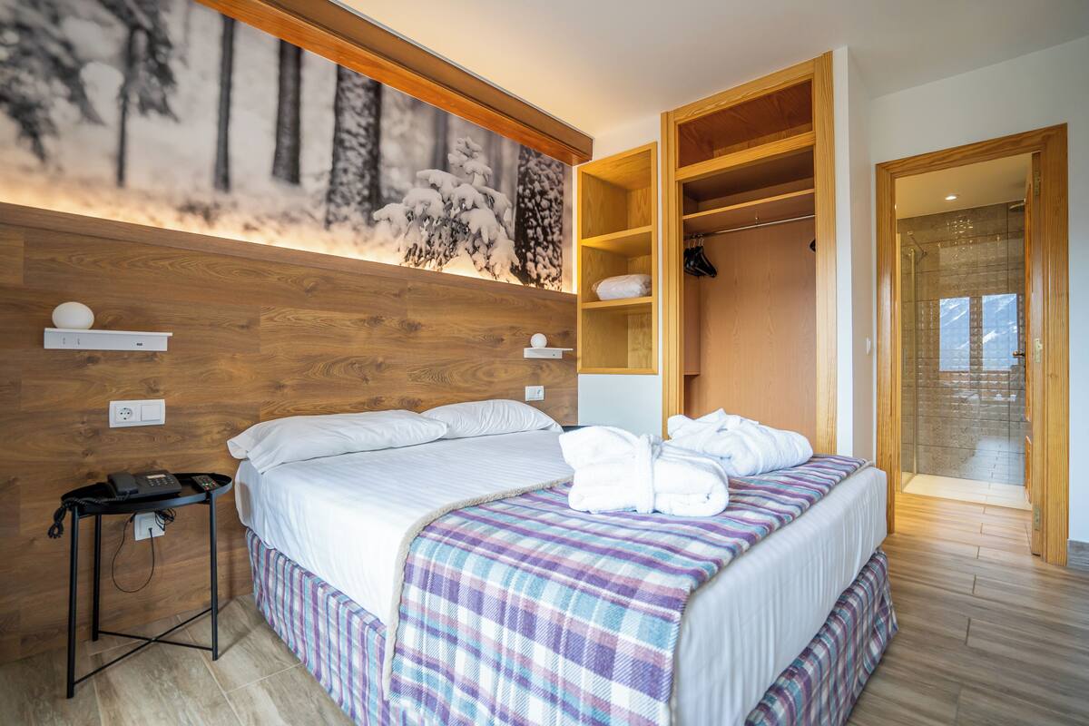 Une chambre d’hôtel moderne avec une tête de lit en bois, un lit recouvert d’une couverture à carreaux, une table de chevet avec un téléphone, un porte-manteau et une armoire.