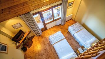 A room with two beds, a wooden ceiling, a window with curtains, a radiator, and a small desk with a television.