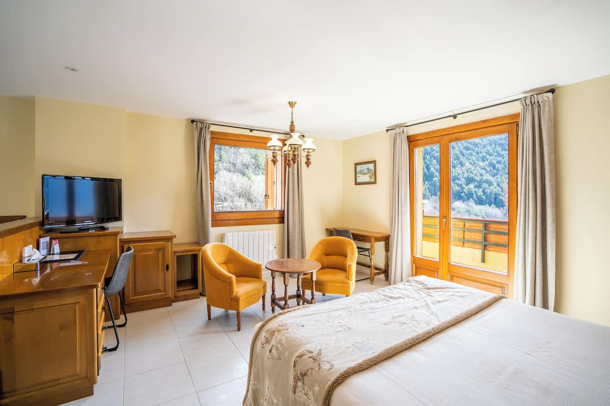 Une chambre d’hôtel équipée d’un lit, d’un bureau, de deux chaises, d’une télévision et de grandes fenêtres donnant sur la verdure.