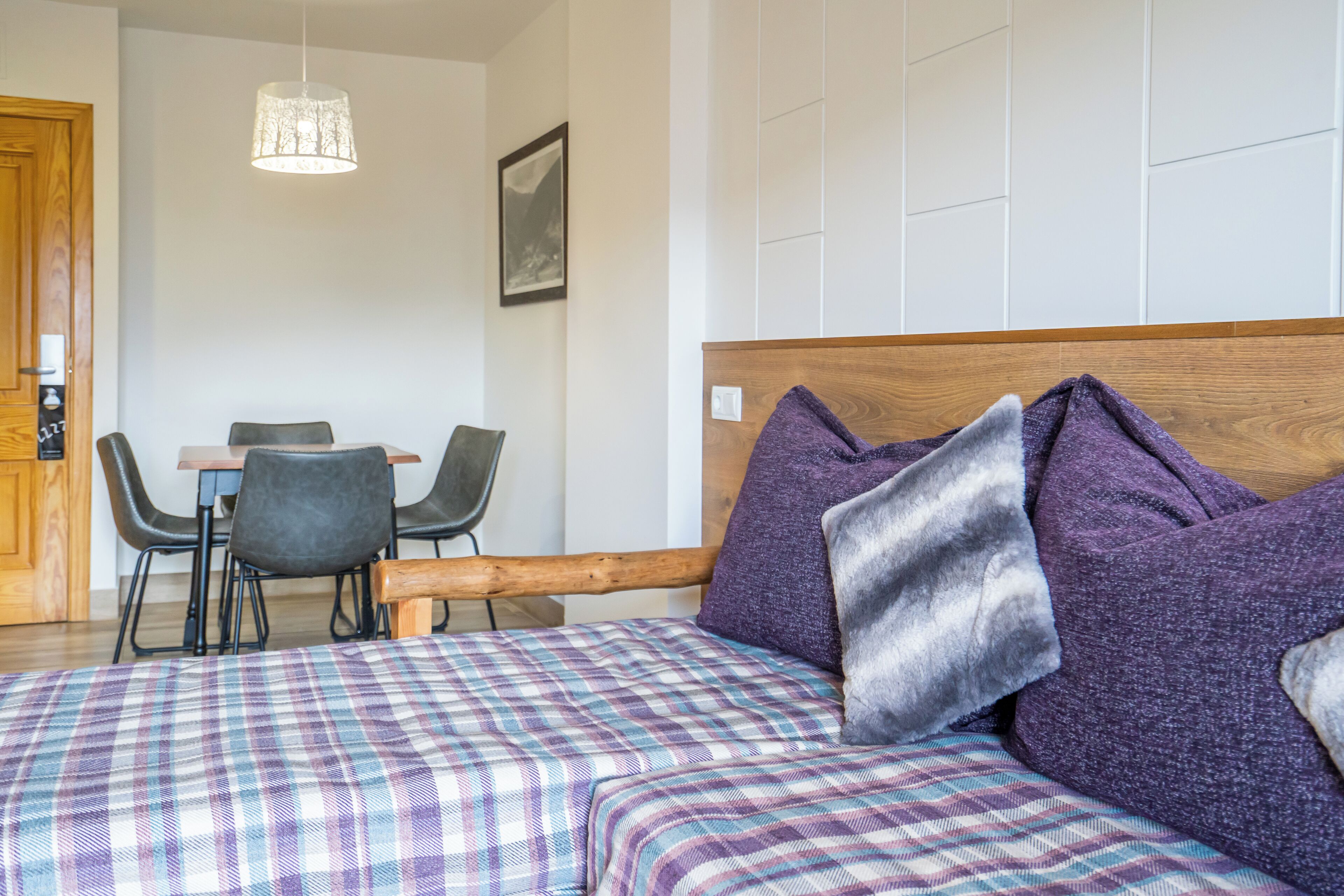 A bedroom with a wooden headboard, plaid bedding, and purple pillows. There is a dining area with chairs and a table in the background.