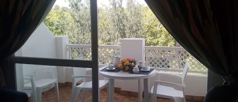 A balcony with white chairs and a table, a white fence, and a view of trees and buildings.