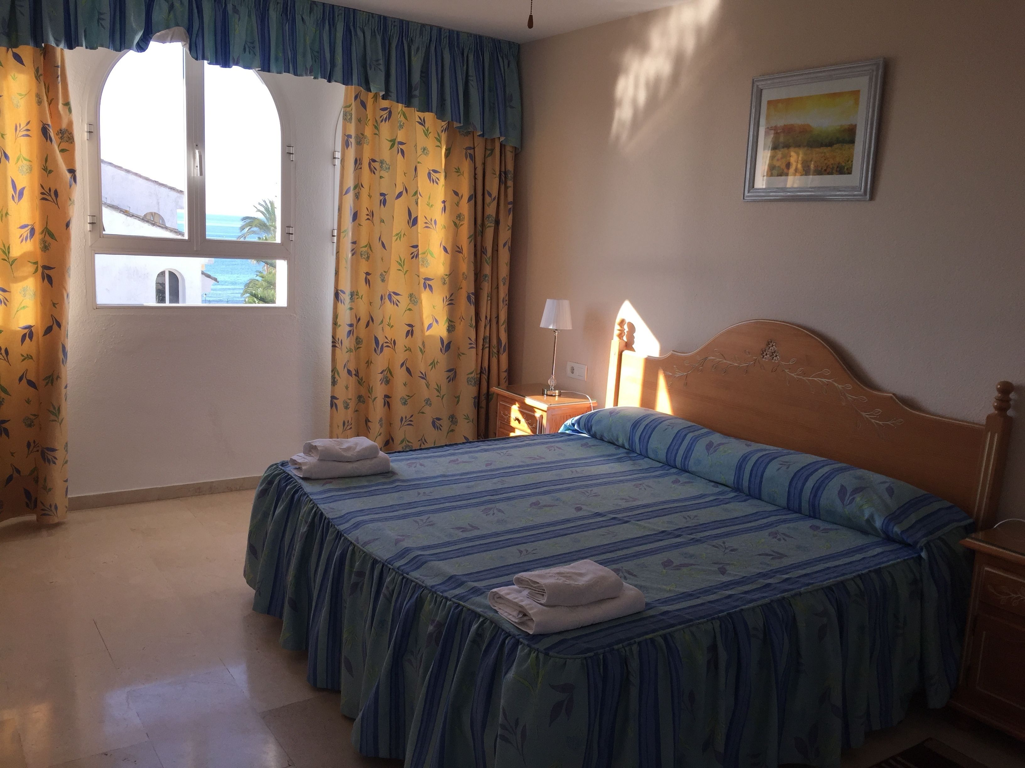 Un dormitorio con cama, cortinas, mesita de noche, lámpara y un cuadro en la pared.