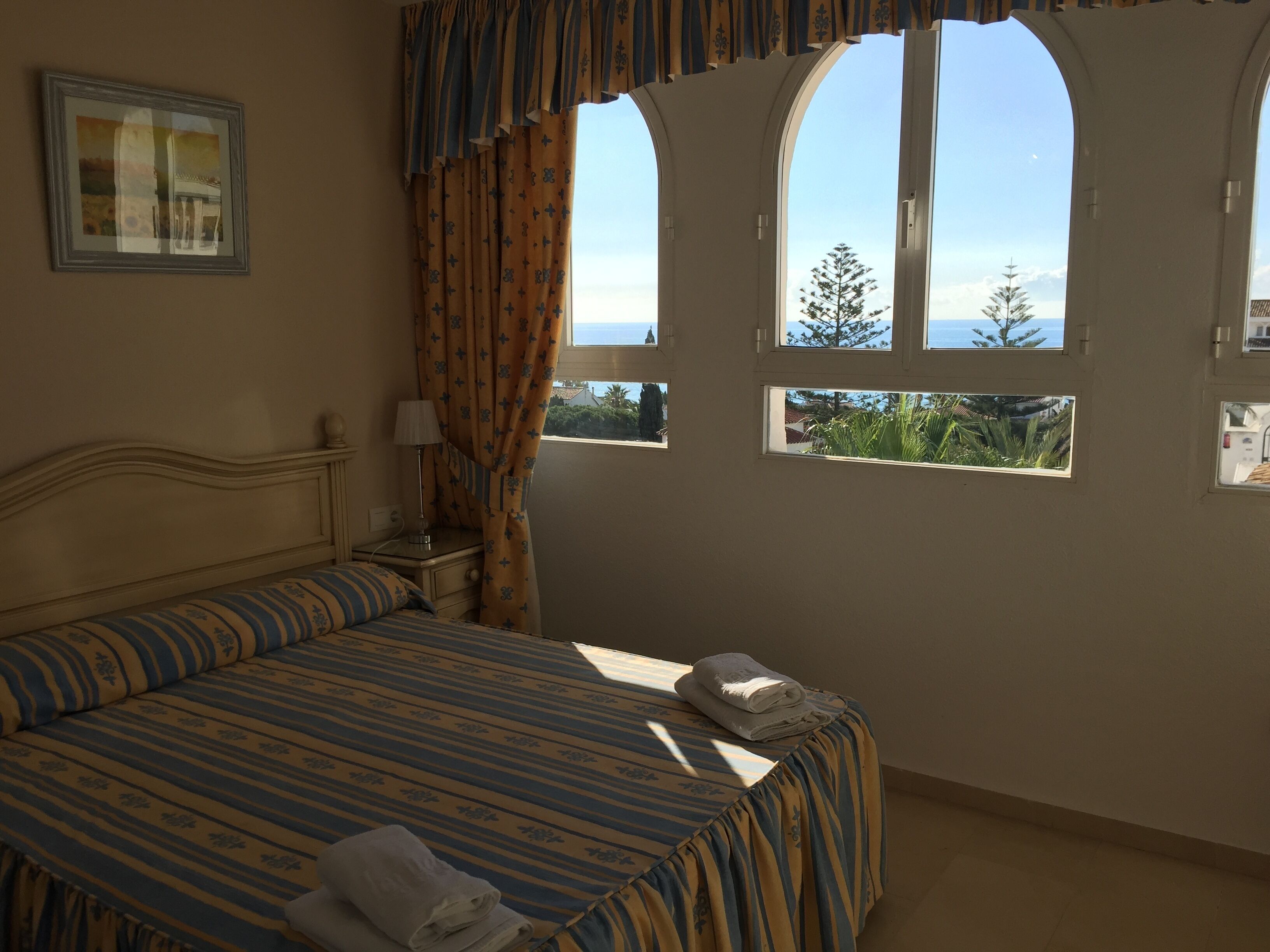 A bedroom with a bed, a nightstand, a lamp, curtains, and a view of the sea through arched windows.