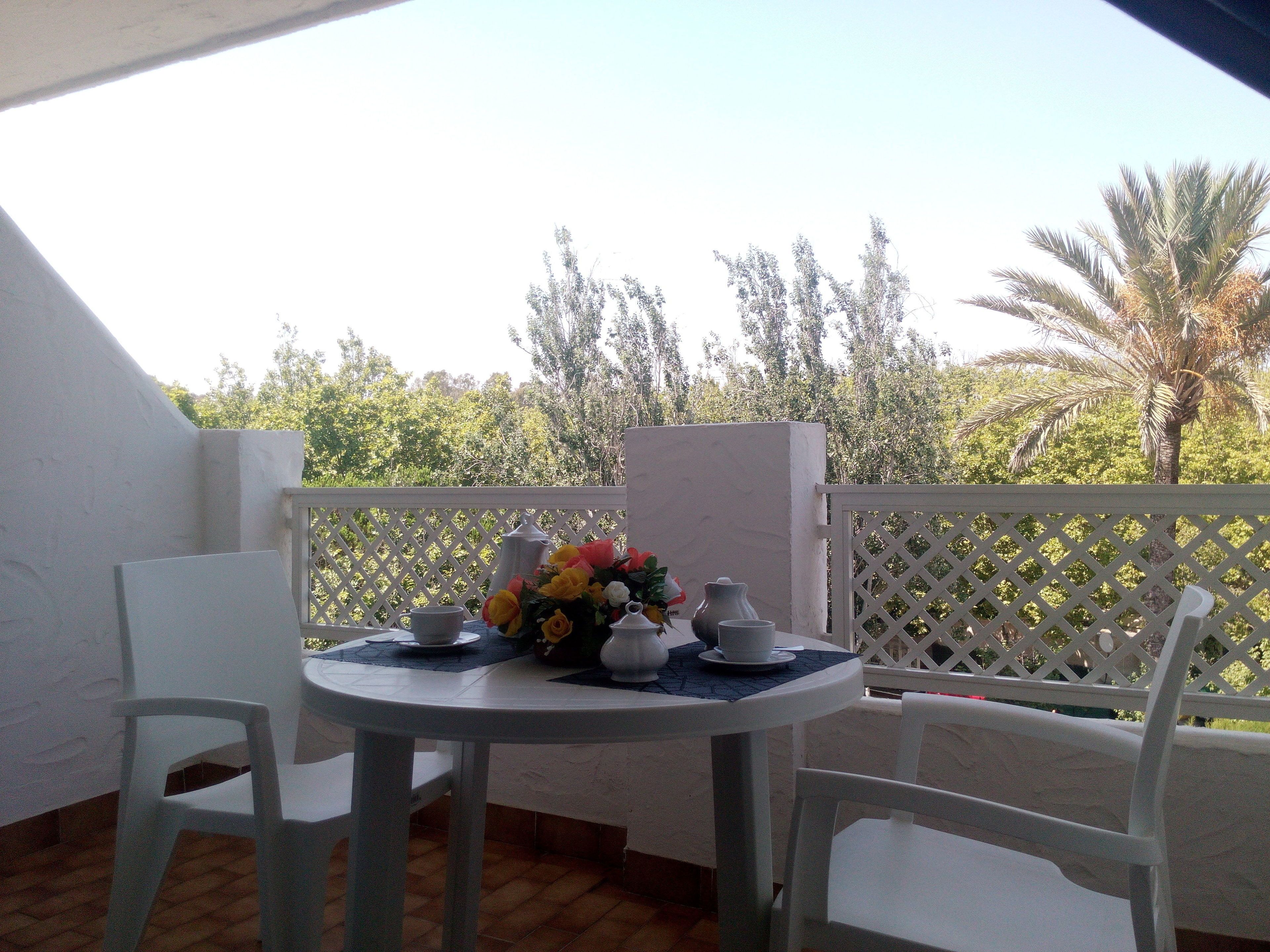 A balcony with a round table set for two, featuring white chairs, a vase of flowers, and a teapot.