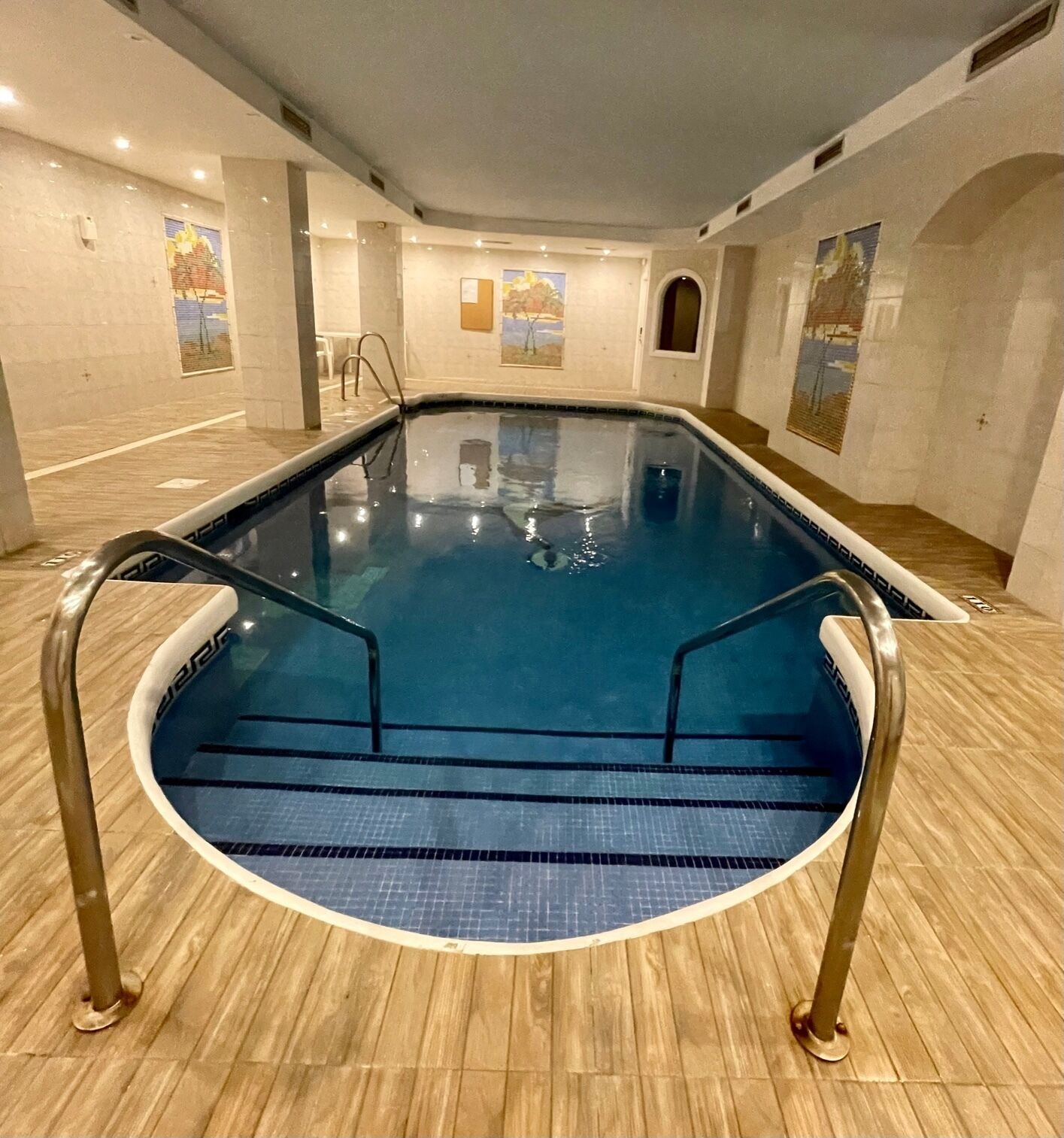 An indoor swimming pool with clear blue water, surrounded by wooden flooring and walls with artwork.