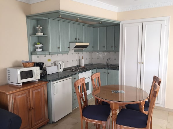 Una cocina compacta con armarios azul verdoso, microondas, tetera y estufa. Hay una mesa de comedor de madera con sillas.