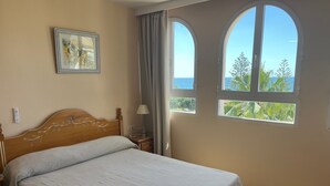 Un lit bien fait, avec une tête de lit en bois, une table de chevet surmontée d’une lampe, un tableau encadré accroché au mur, et de grandes fenêtres en arc offrant une vue sur la mer et la verdure.