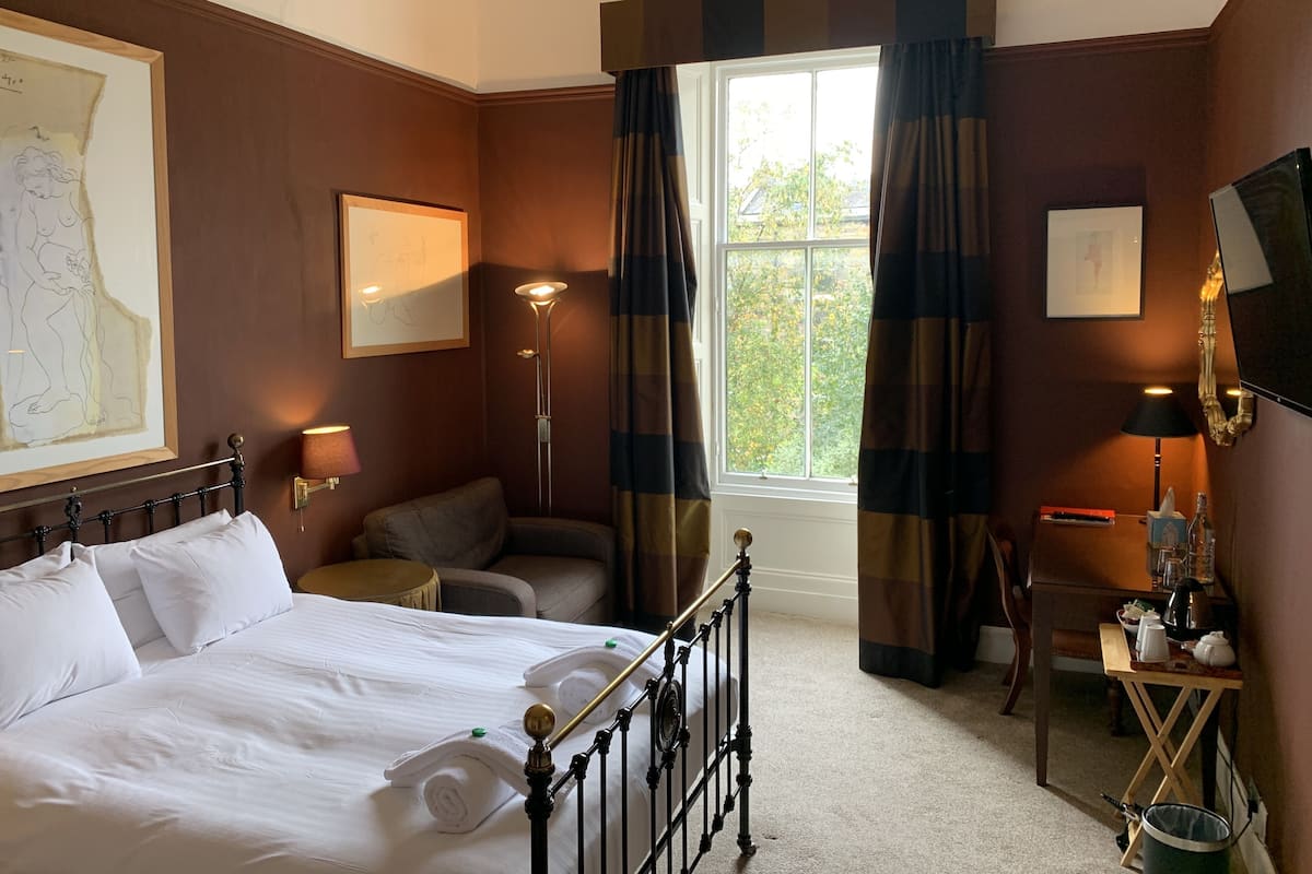 A hotel room with a large bed, a desk with a lamp, a chair, and a window with curtains.