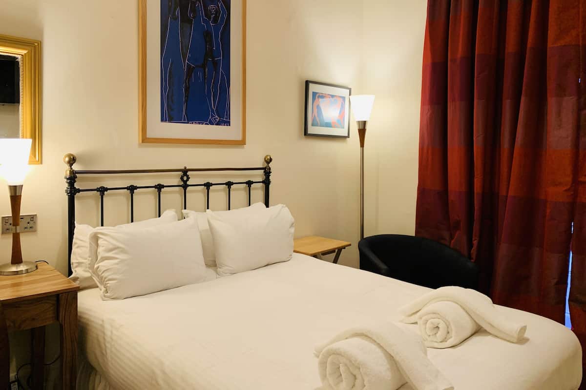A neatly made bed with white linens, a black metal headboard, a framed abstract painting above the bed, a bedside table with a lamp, and a red curtain partially drawn to the side.