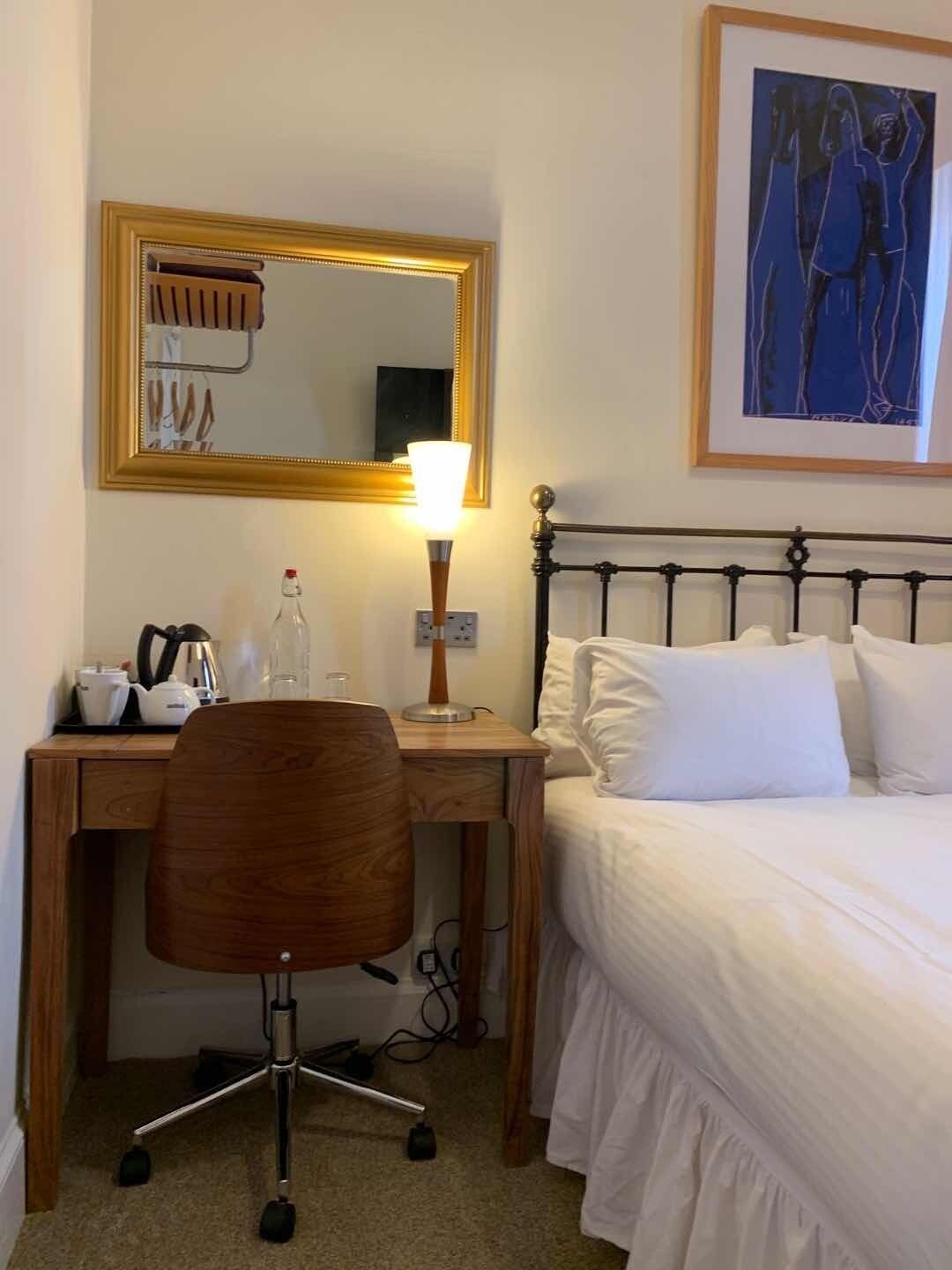 Una habitación de hotel con cama, escritorio con silla, lámpara, espejo y una obra de arte enmarcada en la pared.