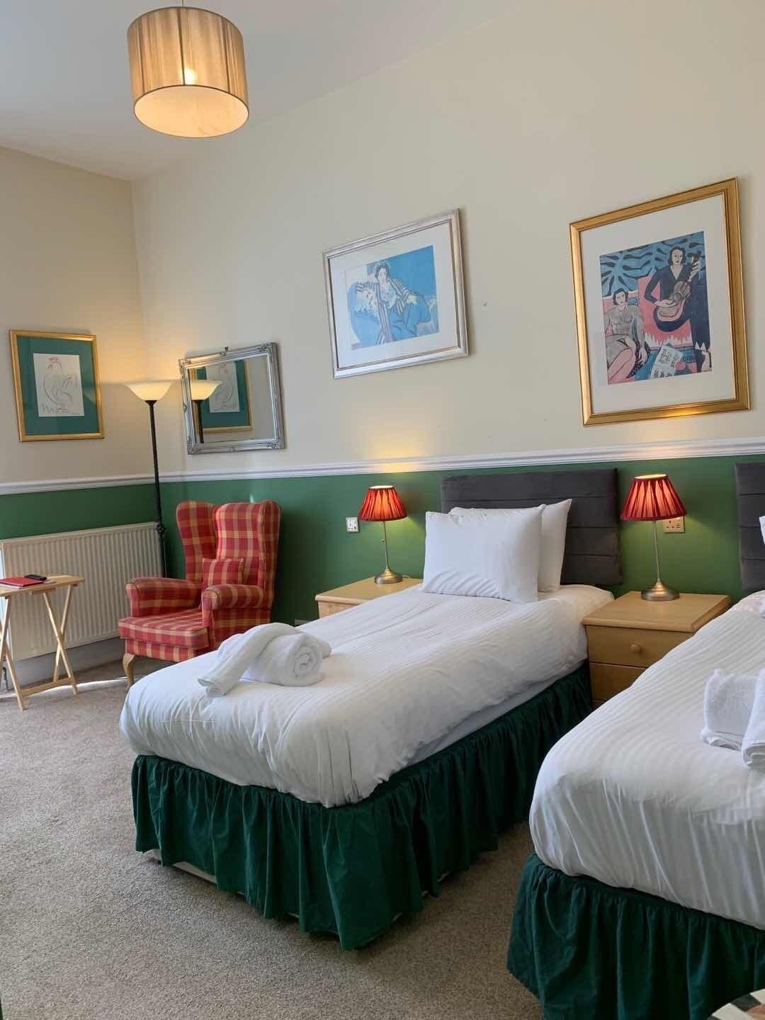 A hotel room with two beds, a chair, a mirror, and artwork on the walls.