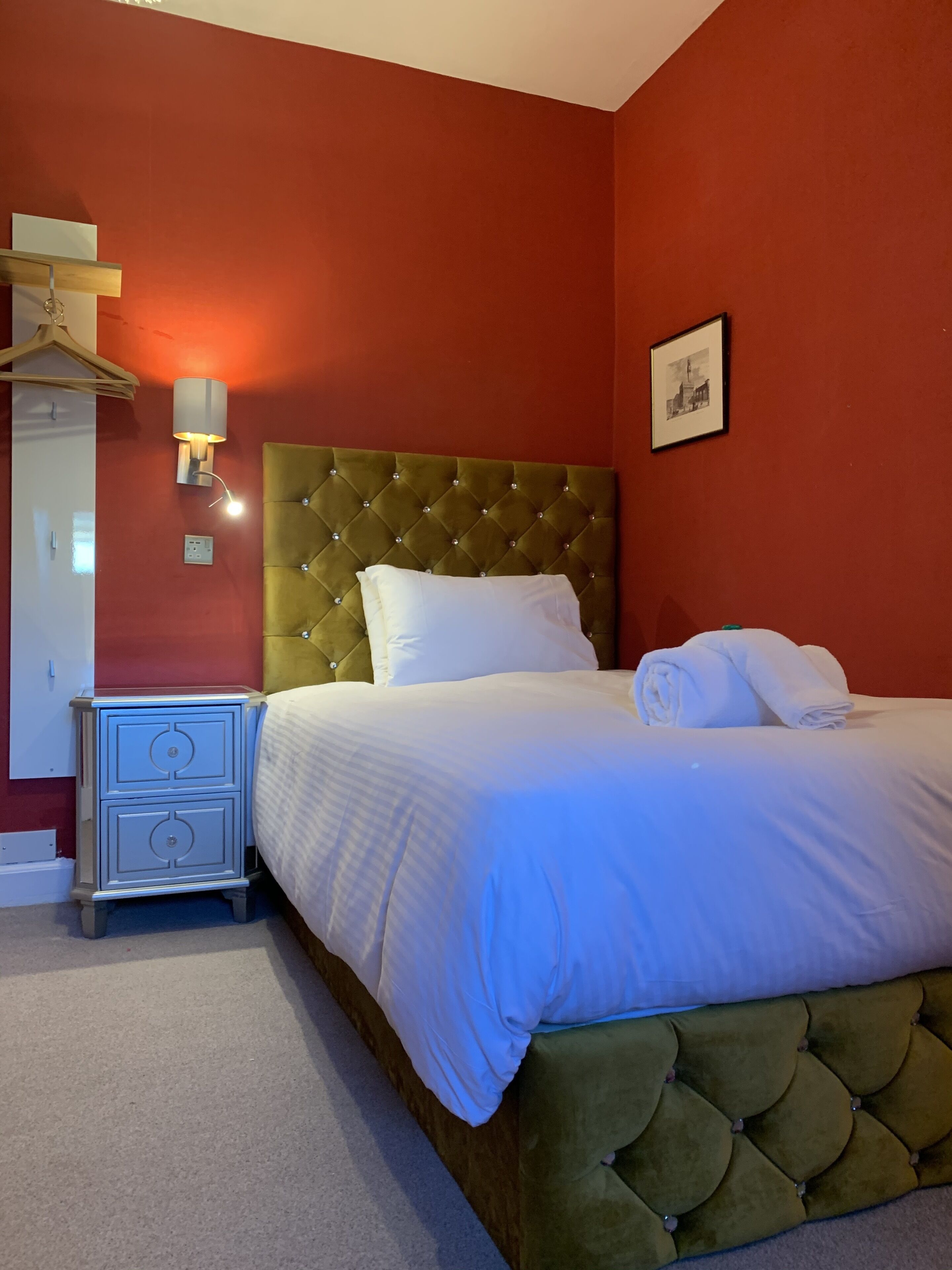 A hotel room with a green tufted headboard, white bedding, a bedside table with drawers, a wall-mounted light, and a framed picture on the wall.