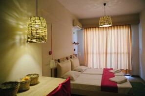A hotel room with two beds, a wooden table, wicker lamps, a red chair, and a window with curtains. - Gorgona Studios (Rhodes)