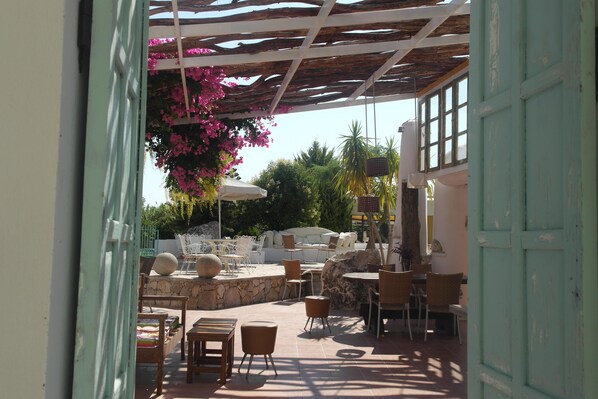 Un espace extérieur aménagé avec des tables et des chaises, une pergola ornée de fleurs roses et une vue sur la verdure.