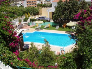 Una piscina rodeada de sillas de descanso y sombrillas, con zonas verdes y edificios residenciales al fondo.