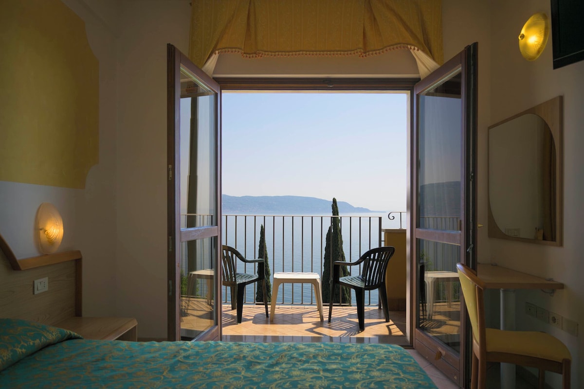 Une chambre avec un lit, une petite table, deux chaises, un balcon avec vue et un grand miroir.