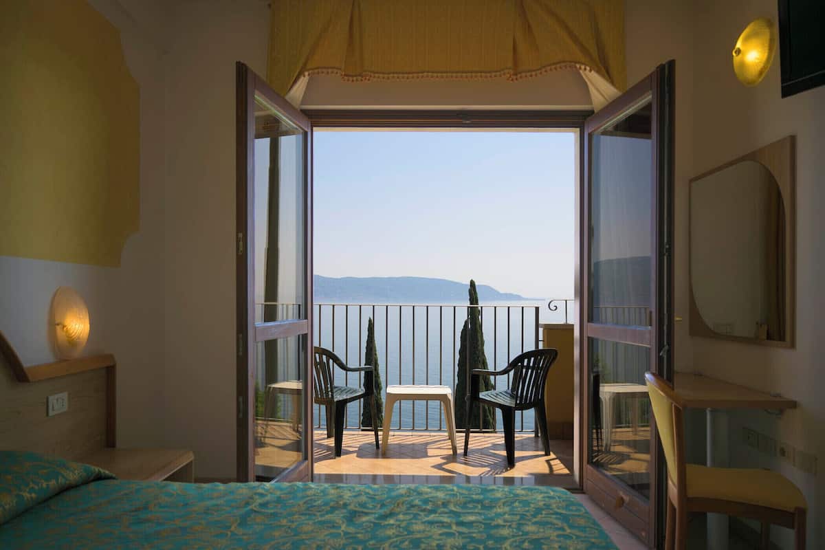 A room with a bed, a small table, two chairs, a balcony with a view, and a large mirror.