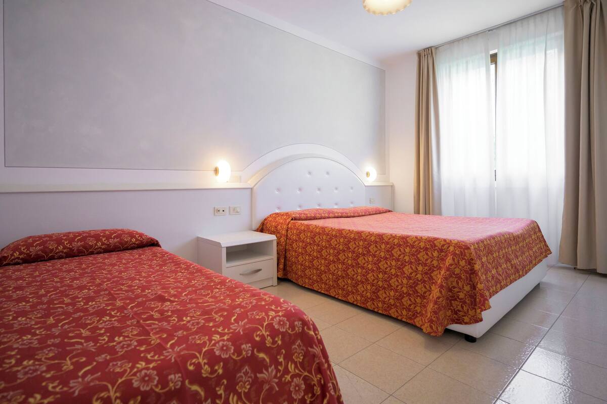 A hotel room with two beds, a white headboard, a nightstand, and a window with curtains.