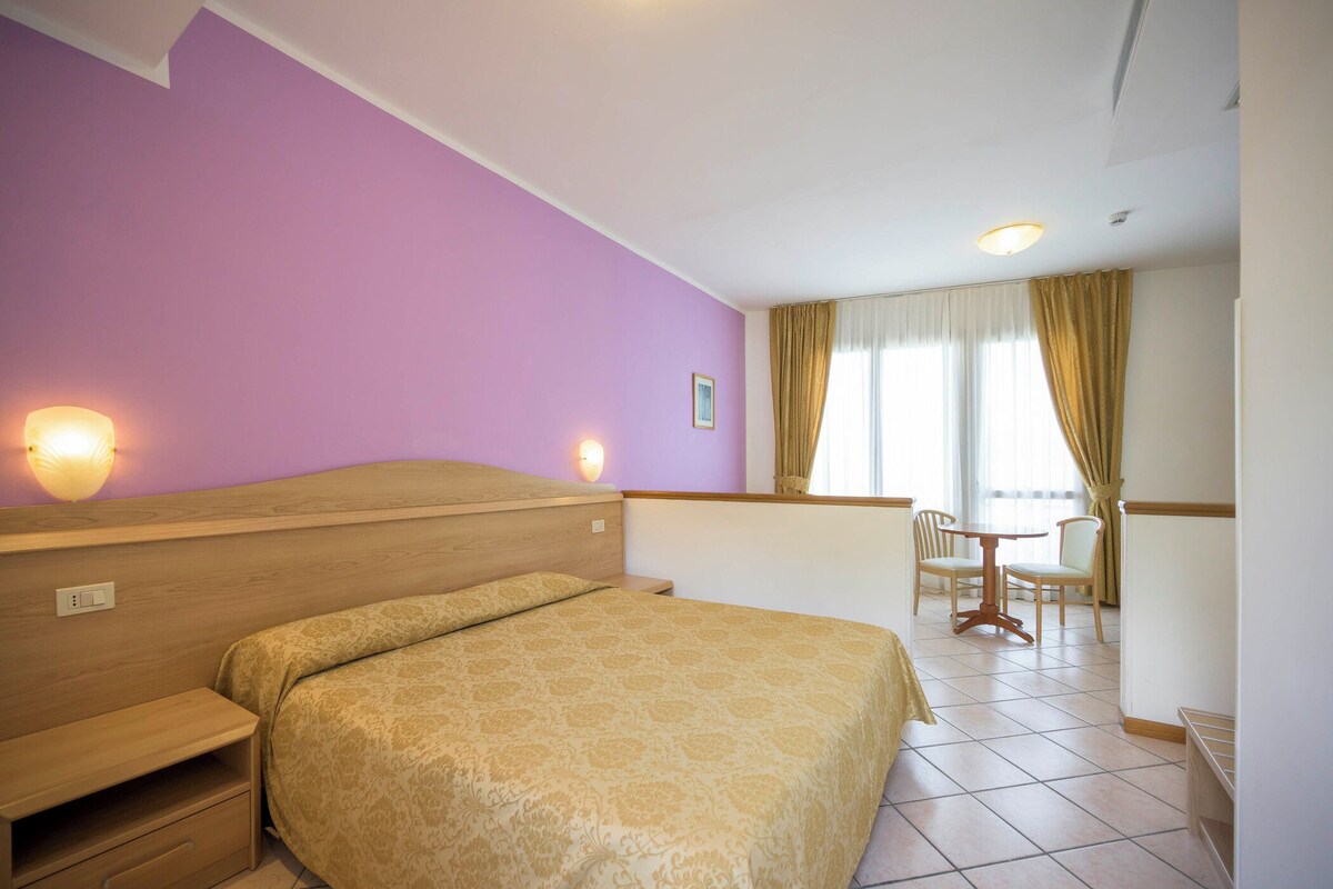 A hotel room with a bed, bedside table, lamp, and a view of a dining area with a table and chairs.