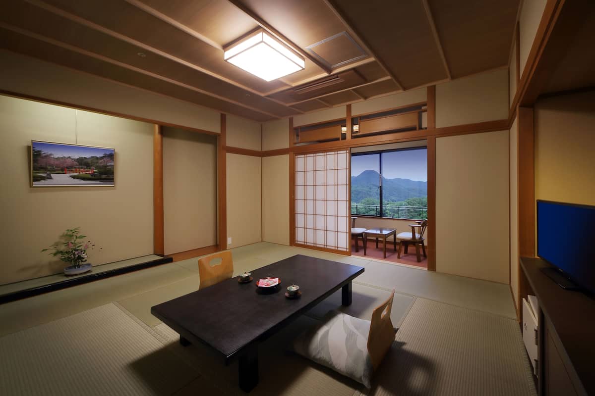 Um quarto japonês tradicional com tatames, uma mesa baixa com almofada, uma televisão e vista para montanhas através de uma porta deslizante.