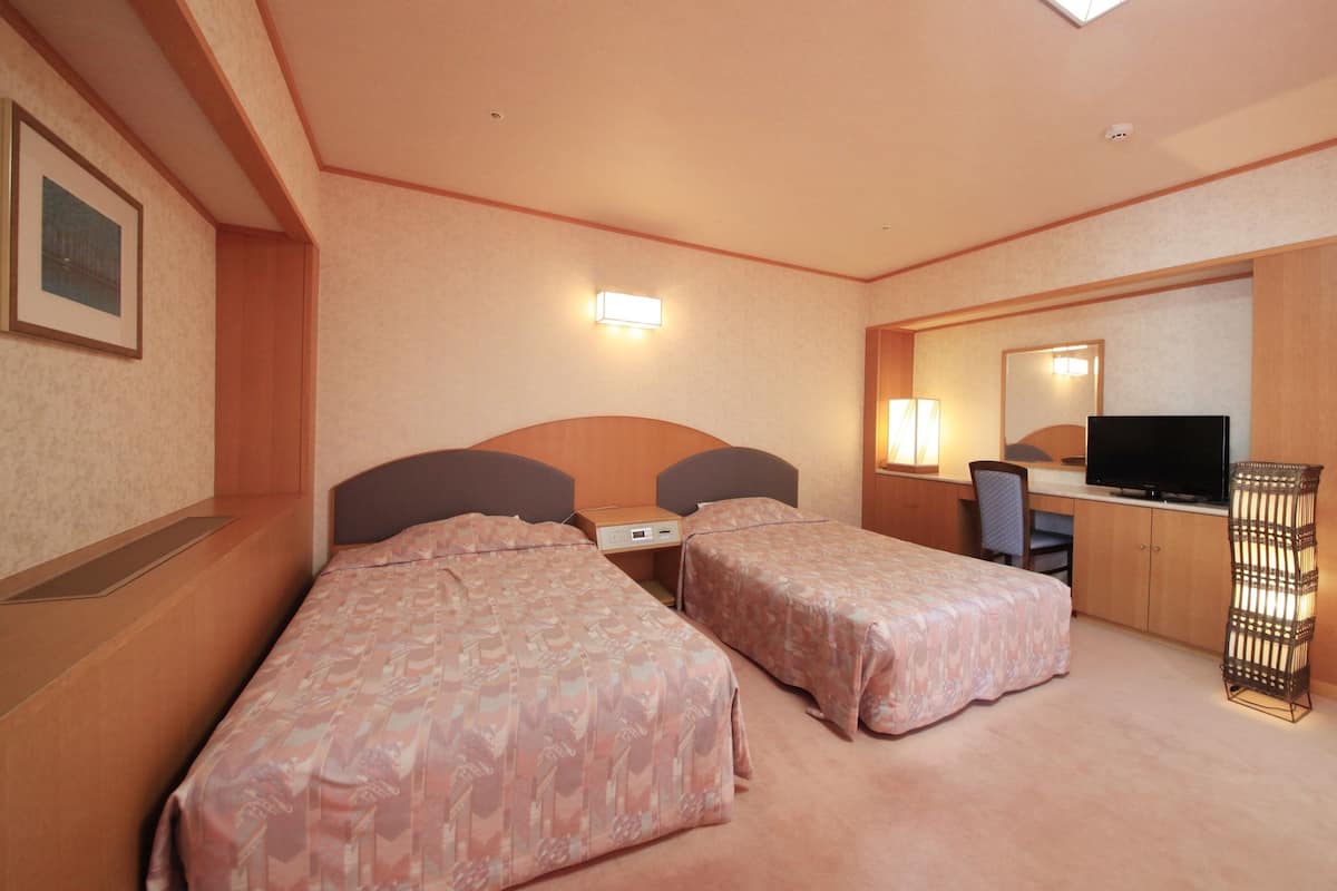 Quarto de hotel com duas camas, uma escrivaninha com computador, televisão e luminária.