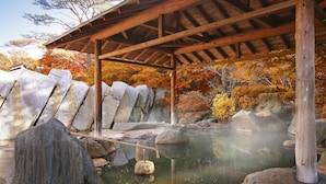Un onsen japonés tradicional con estructura de madera, un baño de piedra y follaje otoñal al fondo.