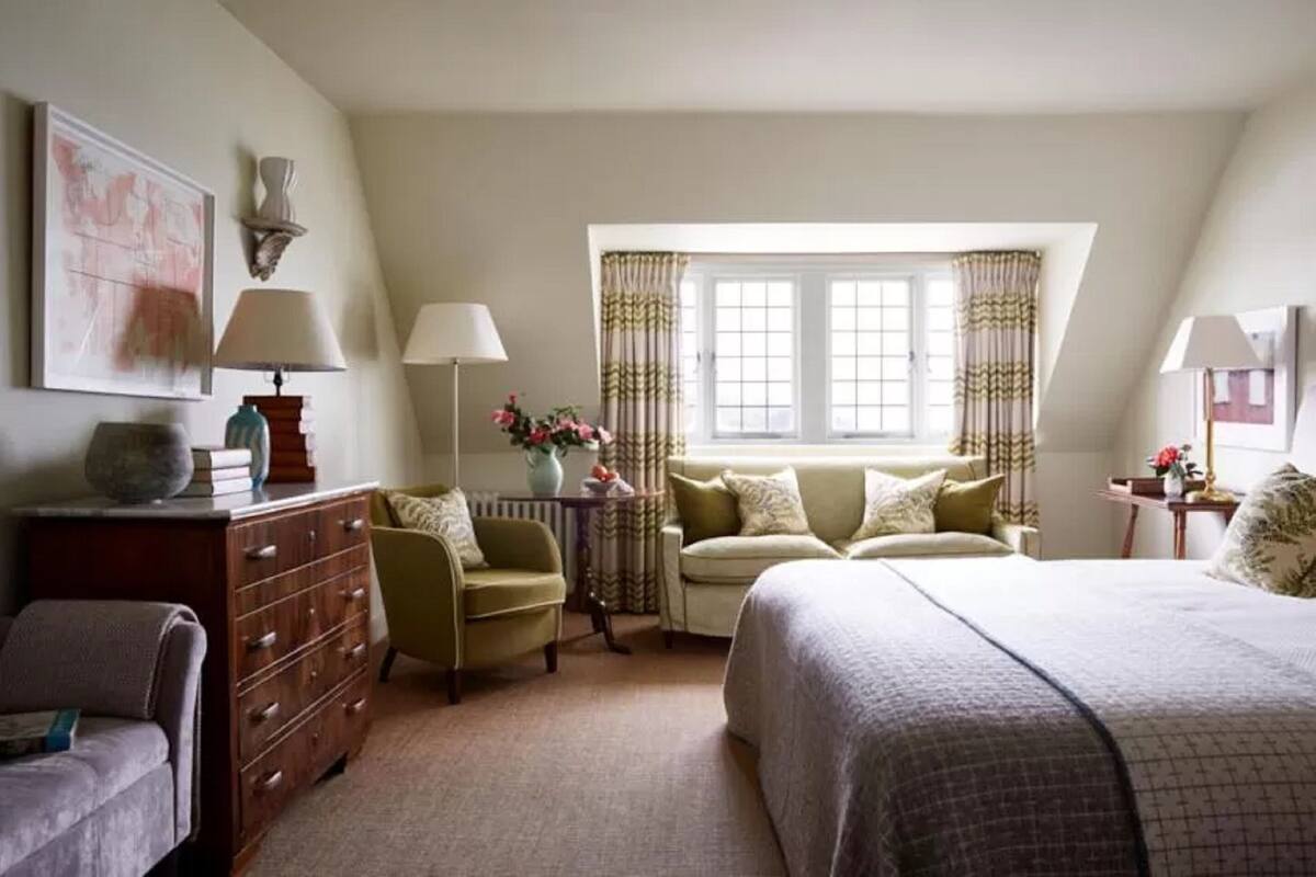 Quarto com cama, duas cadeiras, cômoda, janela com cortinas e abajur.