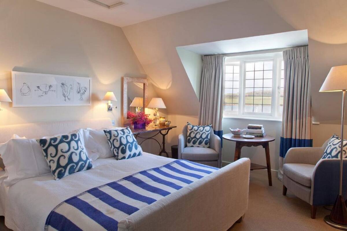 Quarto de hotel com uma cama grande, duas poltronas, uma mesinha e uma janela com cortinas.