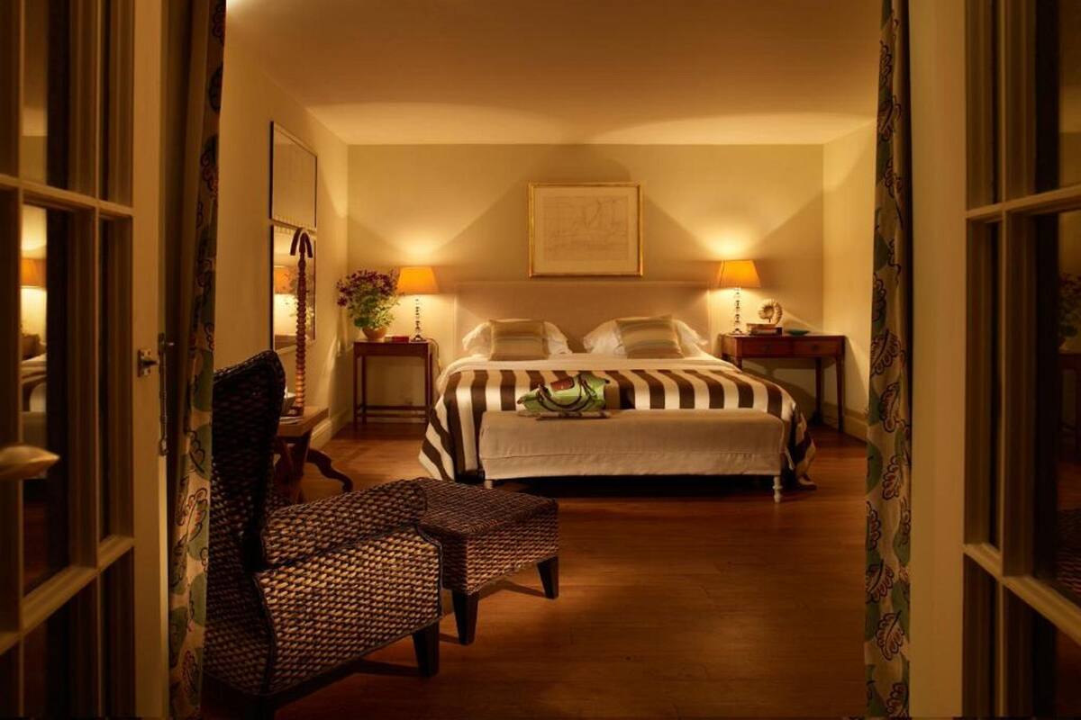 Quarto de hotel com uma cama grande, duas mesas de cabeceira com abajures, uma cadeira e um espelho.