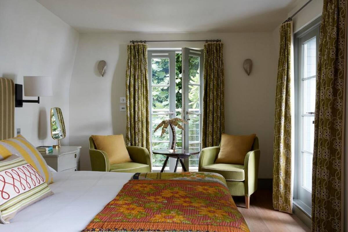 Quarto de hotel com uma cama, duas poltronas, uma mesinha e uma janela com cortinas.