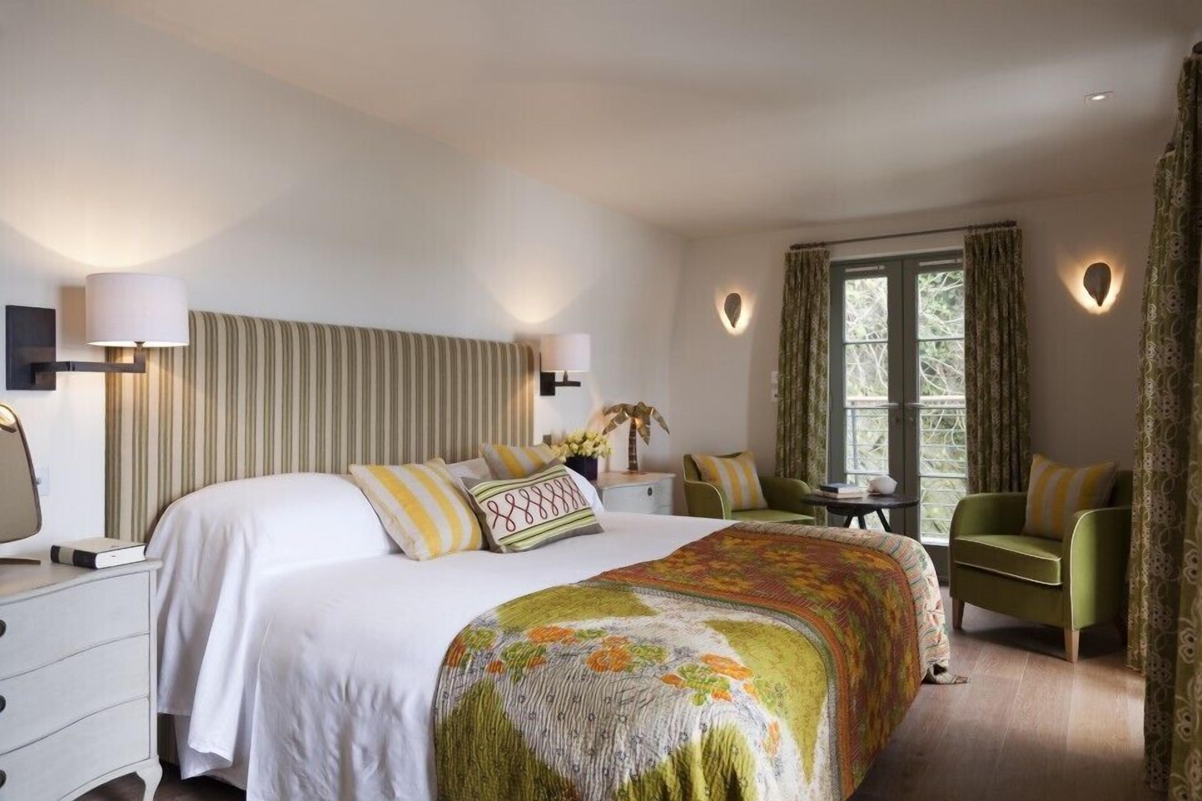 Quarto de hotel com uma cama grande, duas mesas de cabeceira, uma poltrona verde, uma janela com cortinas e um ponto de luz fixado na parede.