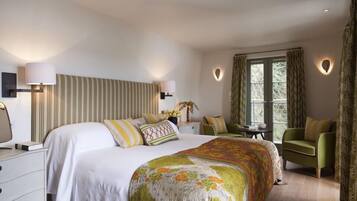Quarto de hotel com uma cama grande, duas mesas de cabeceira, uma poltrona verde, uma janela com cortinas e um ponto de luz fixado na parede.