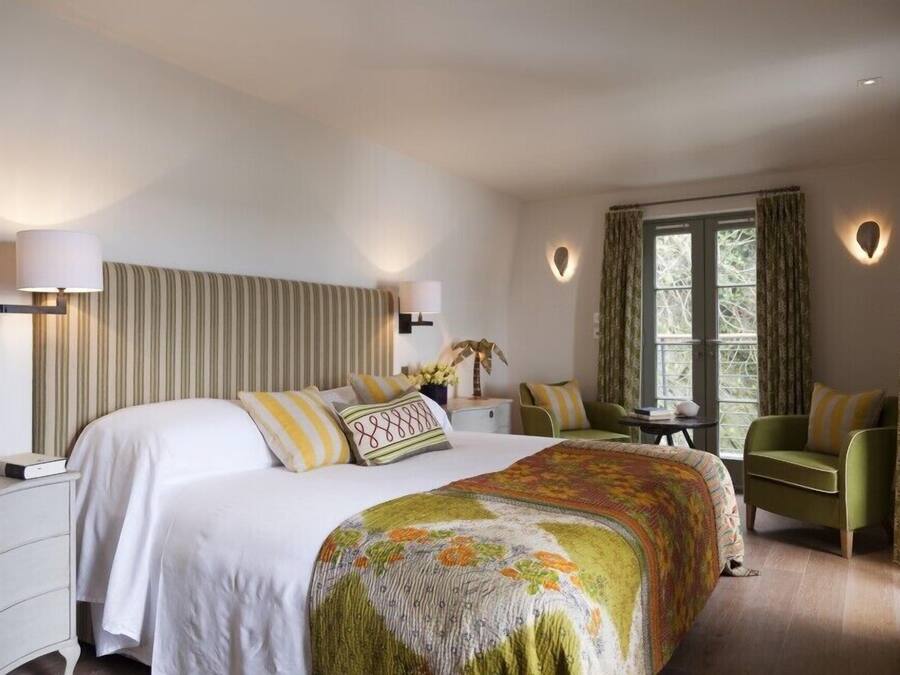Quarto de hotel com uma cama grande, duas mesas de cabeceira, uma poltrona verde, uma janela com cortinas e um ponto de luz fixado na parede.
